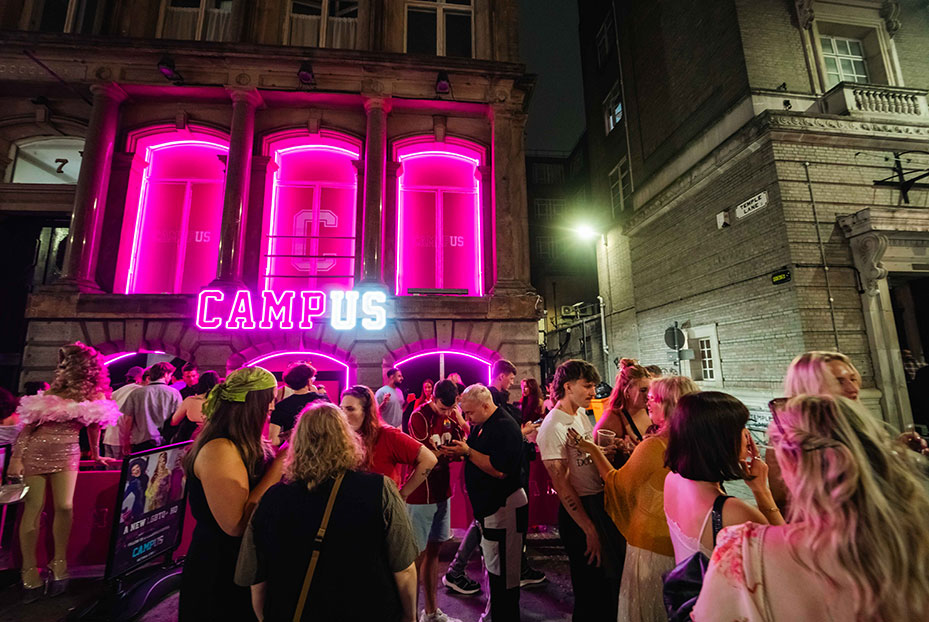 People enjoying drinks and chatting outside Campus bar on Victoria Street at night with neon lights.
